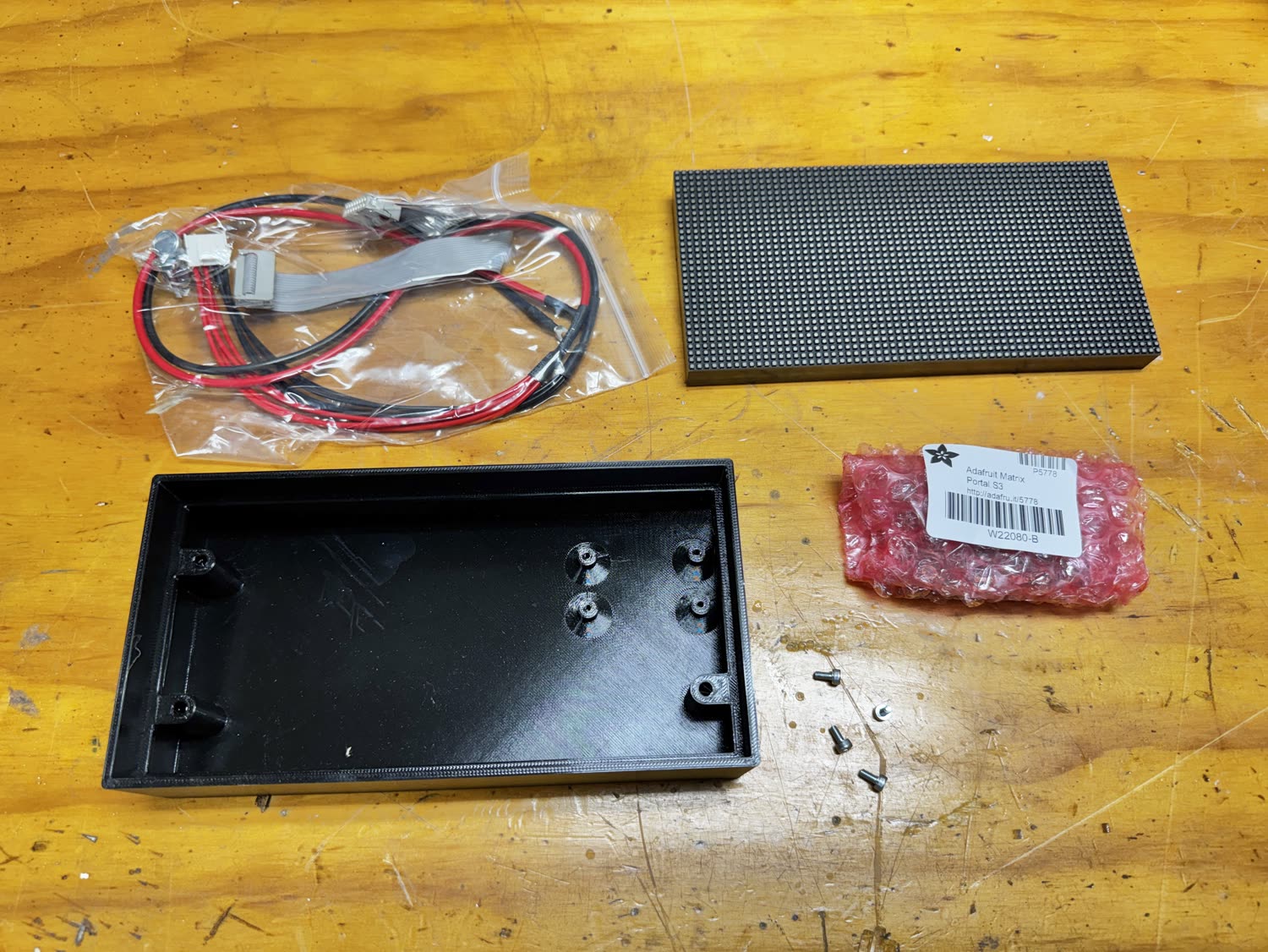A MatrixPortal S3, 64 by 32 LED panel, HUB75 ribbon cable, power leads, screws, and an optional printed enclosure laid out on a workbench