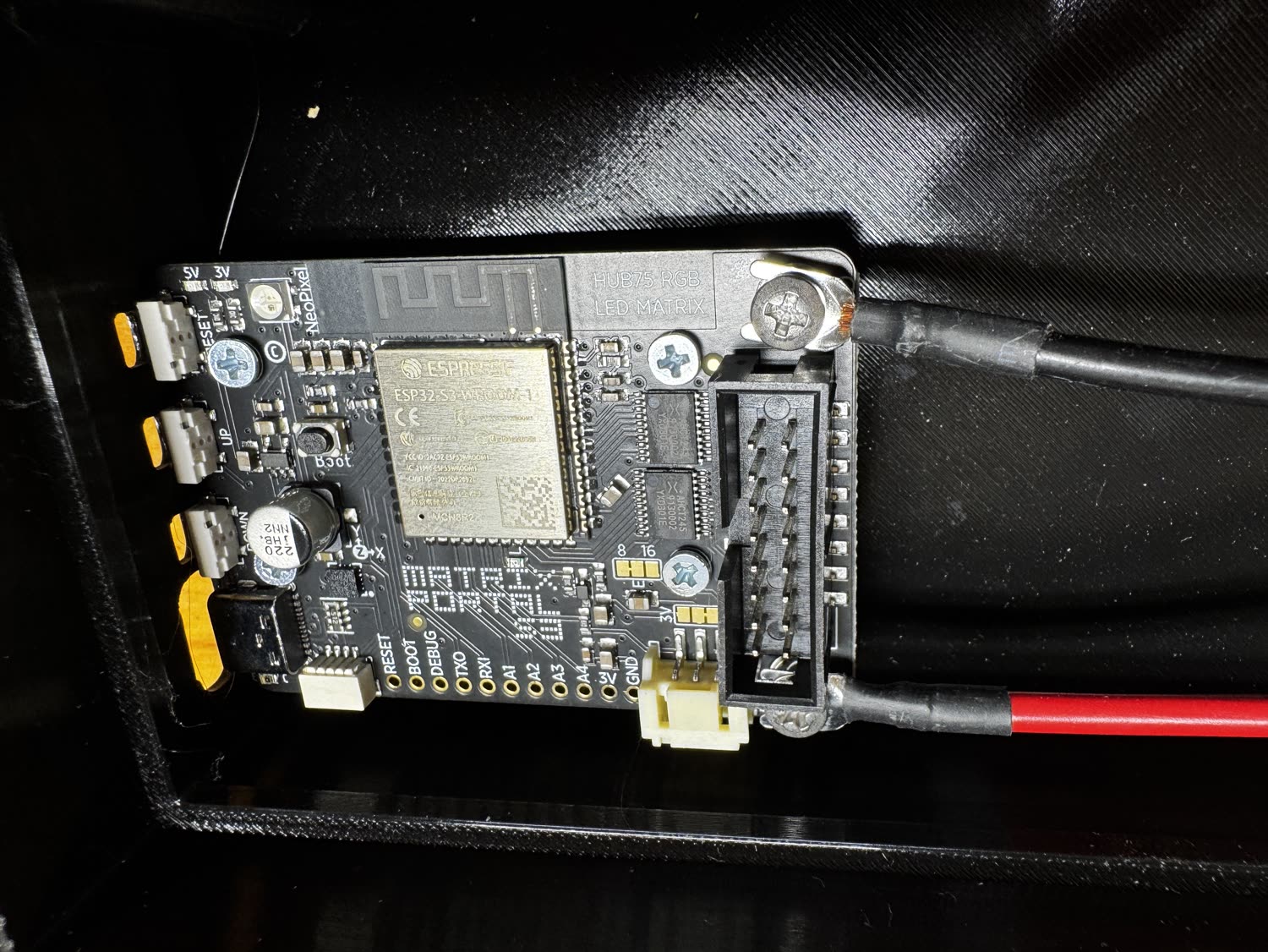 A MatrixPortal S3 with the gray HUB75 ribbon cable connected, black lead on GND, and red lead on 5V